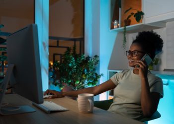 Positive multiracial woman in glasses talking on smartphone, working on desktop pc late at night. Photo credit: @DimaBerlin via Twenty20