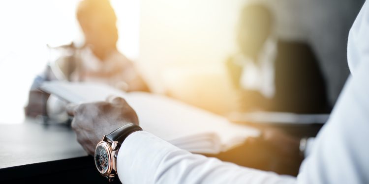 Close-up hand of a businessman in a white shirt with a clock holding documents. In the background are blurry colleagues and bright sunlight